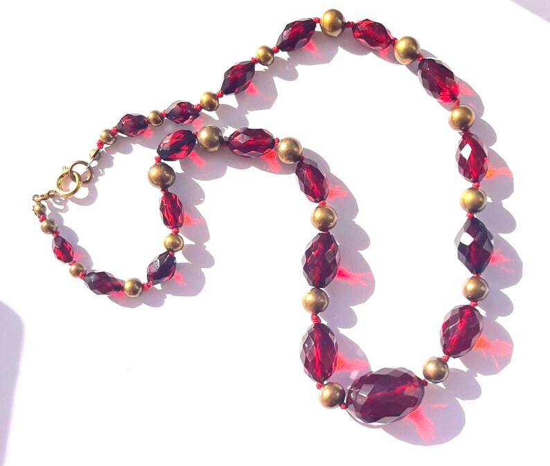 May include: A vintage necklace featuring faceted, deep red glass beads and small gold-toned beads. The necklace has a lobster clasp closure. The beads are strung on a red cord. The necklace is displayed on a white surface.