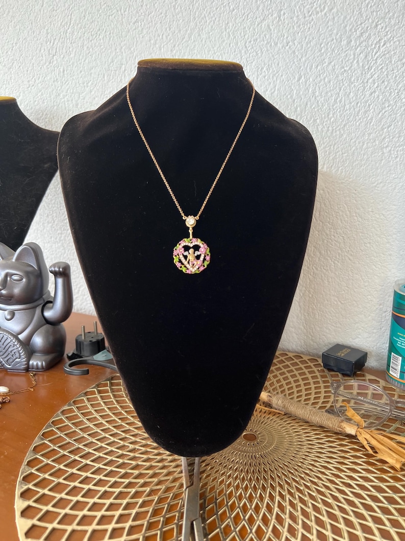 K&ouml;nnte beinhalten: Eine goldene Kettenkette mit einem herzf&ouml;rmigen Anh&auml;nger. Der Anh&auml;nger ist mit rosa und gr&uuml;nen Kristallen verziert und zeigt eine kleine Engelsfigur im Inneren.