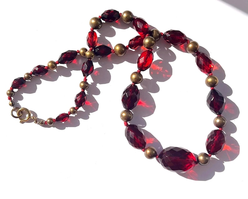 May include: A vintage necklace featuring faceted, deep red beads interspersed with small, round, gold-toned beads. The necklace has a classic design with a gold-colored clasp. The beads are arranged on a red string, creating a striking contrast.