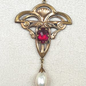 Op de afbeelding: Een bronzen metalen ketting met een hanger die een ingewikkeld opengewerkt frame heeft met een centrale ovaal geslepen dieprode edelsteen. Een enkele druppelvormige witte parel hangt aan de basis van de metalen zetting aan een bijpassende ketting.