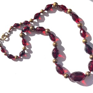 May include: A vintage necklace featuring faceted, deep red glass beads and small, round gold-tone beads. The necklace is strung on red thread and has a gold-tone clasp. The beads are arranged in a repeating pattern, creating a classic and elegant design.