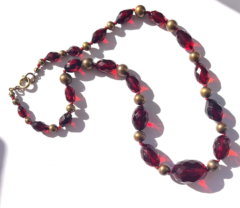May include: A vintage necklace featuring faceted, deep red glass beads and small, round gold-tone beads. The necklace is strung on red thread and has a gold-tone clasp. The beads are arranged in a repeating pattern, creating a classic and elegant design.
