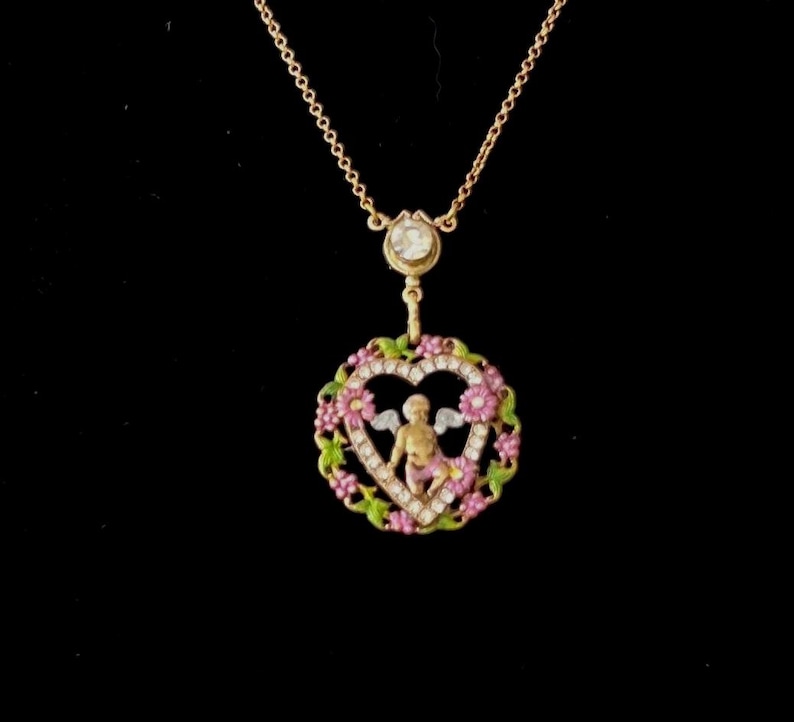 K&ouml;nnte beinhalten: Eine goldene Halskette mit einem herzf&ouml;rmigen Anh&auml;nger, der einen Cherub in einem Kranz aus rosa und gr&uuml;nen Blumen zeigt. Der Anh&auml;nger ist mit kleinen wei&szlig;en Steinen verziert.