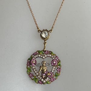 K&ouml;nnte beinhalten: Eine goldene Halskette mit einem herzf&ouml;rmigen Anh&auml;nger, der einen Cherub mit Fl&uuml;geln in einem Blumengekr&auml;nz zeigt. Der Anh&auml;nger ist mit klaren Strasssteinen und rosa und gr&uuml;nem Email verziert.