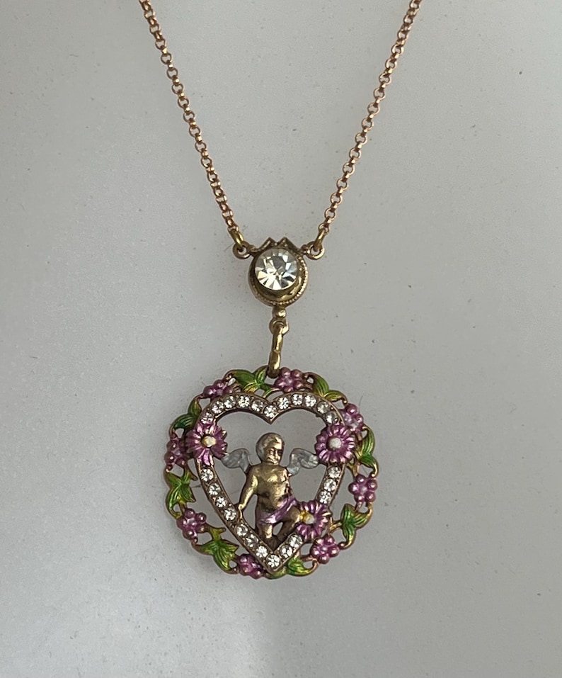 K&ouml;nnte beinhalten: Eine goldene Halskette mit einem herzf&ouml;rmigen Anh&auml;nger, der einen Cherub mit Fl&uuml;geln in einem Blumengekr&auml;nz zeigt. Der Anh&auml;nger ist mit klaren Strasssteinen und rosa und gr&uuml;nem Email verziert.