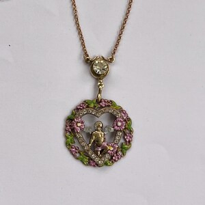 K&ouml;nnte beinhalten: Eine goldene Halskette mit einem herzf&ouml;rmigen Anh&auml;nger, der einen Cherub mit Fl&uuml;geln im Inneren zeigt. Das Herz ist von rosa und gr&uuml;nen Emaille-Blumen und Strasssteinen umgeben.