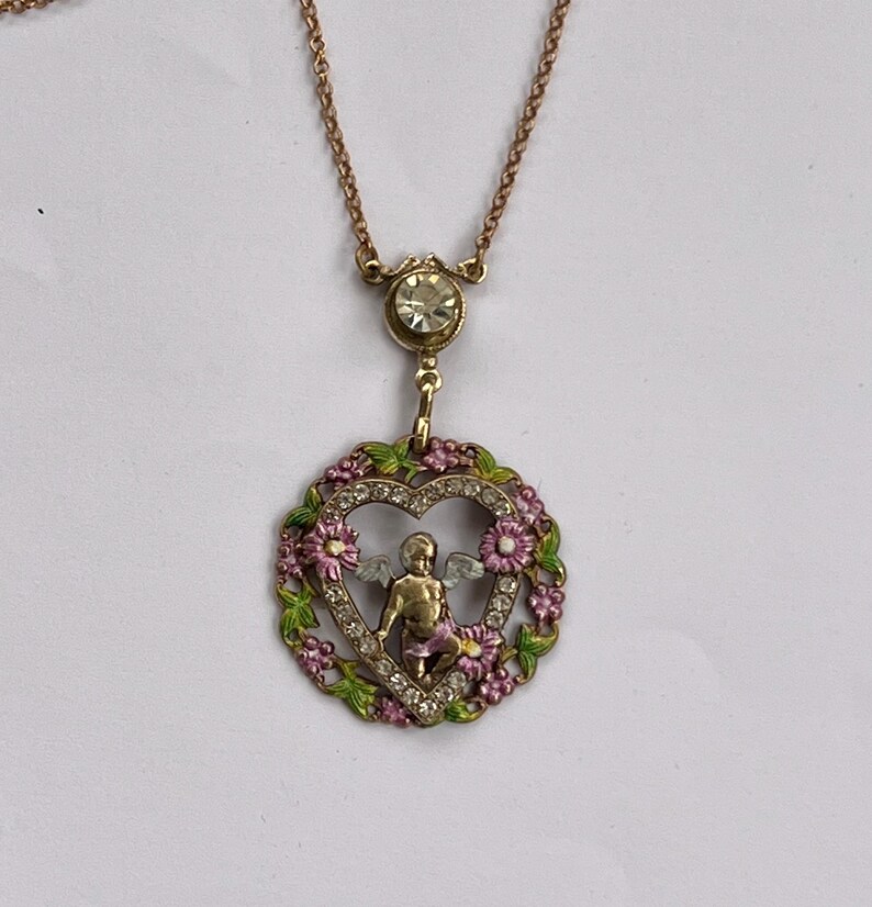K&ouml;nnte beinhalten: Eine goldene Halskette mit einem herzf&ouml;rmigen Anh&auml;nger, der einen Cherub mit Fl&uuml;geln im Inneren zeigt. Das Herz ist von rosa und gr&uuml;nen Emaille-Blumen und Strasssteinen umgeben.