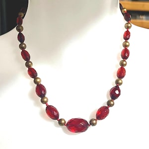 May include: A red beaded necklace with faceted oval and round beads. The necklace features alternating red and gold-colored beads, strung on a red cord. The necklace is displayed on a white mannequin.
