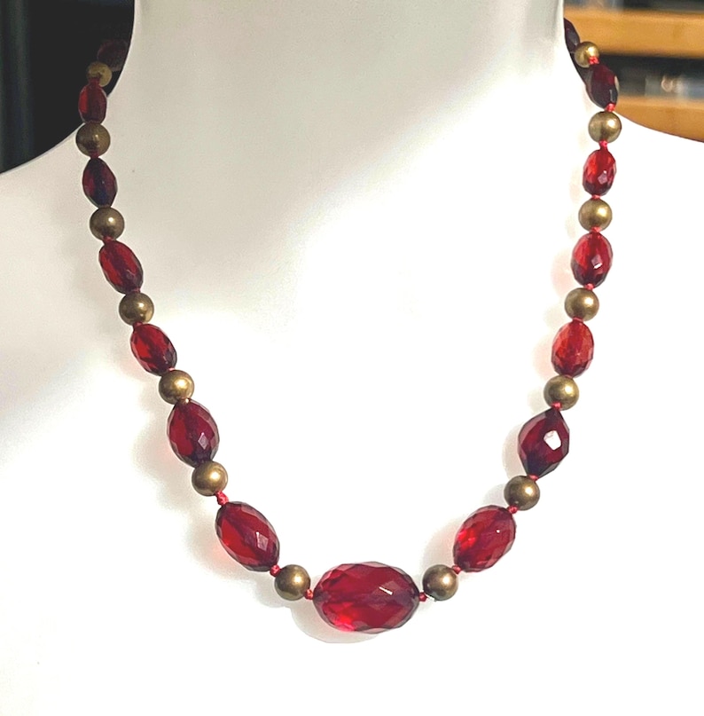 May include: A red beaded necklace with faceted oval and round beads. The necklace features alternating red and gold-colored beads, strung on a red cord. The necklace is displayed on a white mannequin.