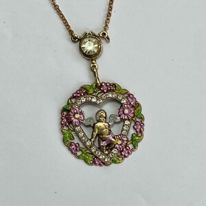 K&ouml;nnte beinhalten: Eine goldfarbene Halskette mit einem herzf&ouml;rmigen Anh&auml;nger, der einen Cherub mit Fl&uuml;geln in einem herzf&ouml;rmigen Rahmen aus rosa und gr&uuml;nem Emaille und Strasssteinen zeigt.