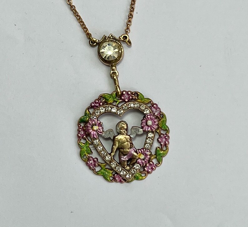 K&ouml;nnte beinhalten: Eine goldfarbene Halskette mit einem herzf&ouml;rmigen Anh&auml;nger, der einen Cherub mit Fl&uuml;geln in einem herzf&ouml;rmigen Rahmen aus rosa und gr&uuml;nem Emaille und Strasssteinen zeigt.