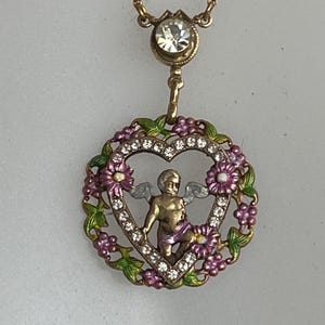 K&ouml;nnte beinhalten: Eine goldfarbene Halskette mit einem herzf&ouml;rmigen Anh&auml;nger, der einen Cherub mit Fl&uuml;geln zeigt, umgeben von rosa und gr&uuml;nen Emaille-Blumen und klaren Strasssteinen. Der Anh&auml;nger h&auml;ngt an einer zarten Kette mit einem klaren Strassstein.