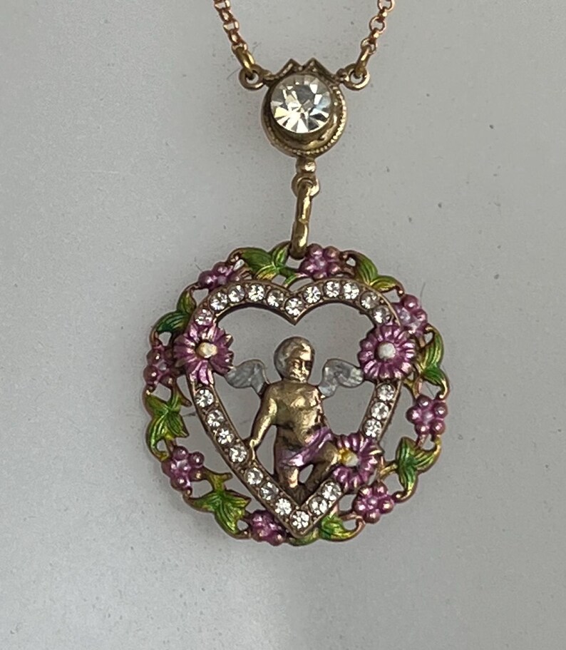 K&ouml;nnte beinhalten: Eine goldfarbene Halskette mit einem herzf&ouml;rmigen Anh&auml;nger, der einen Cherub mit Fl&uuml;geln zeigt, umgeben von rosa und gr&uuml;nen Emaille-Blumen und klaren Strasssteinen. Der Anh&auml;nger h&auml;ngt an einer zarten Kette mit einem klaren Strassstein.