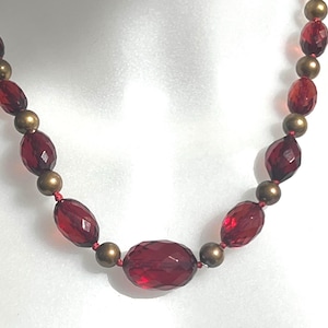 May include: A necklace featuring faceted, oval-shaped, deep red beads interspersed with smaller, round, gold-colored beads. The necklace is strung on a red cord, creating a striking contrast. The beads have a glossy finish.