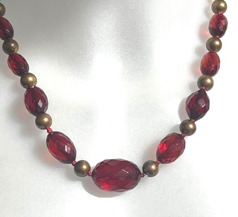 May include: A necklace featuring faceted, oval-shaped, deep red beads interspersed with smaller, round, gold-colored beads. The necklace is strung on a red cord, creating a striking contrast. The beads have a glossy finish.