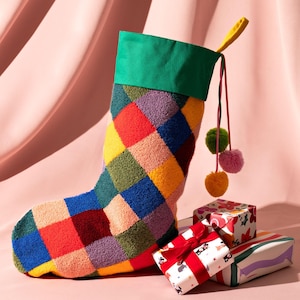 Könnte beinhalten: Ein großer, farbenfroher Weihnachtsstrumpf mit einem Patchwork-Design in verschiedenen Farben, darunter Rot, Blau, Gelb und Grün. Der Strumpf hat eine grüne Manschette und ist mit Pompons verziert. Mehrere verpackte Geschenke befinden sich am Fuß des Strumpfes.
