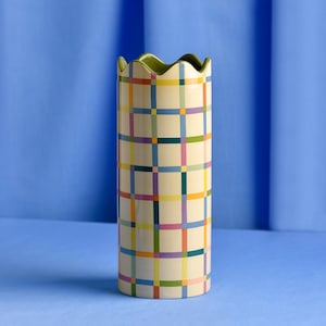 May include: A tall, cylindrical vase with a cream base and a colourful plaid pattern in blue, yellow, orange, green, and purple. The rim has a wavy edge and a green interior. A decorative home accent.