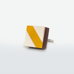 May include: A square earring with a wooden edge, a cream-coloured face, and a diagonal yellow stripe. The earring post is silver. The earring is a geometric design.