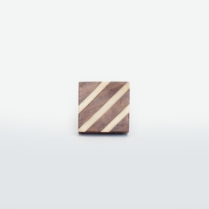 May include: A square, wooden coaster with a dark brown base and three diagonal, light-colored stripes. The coaster is set against a plain white background. The design is simple and geometric.