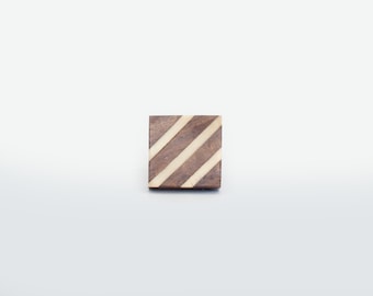 Op de afbeelding: Een vierkante houten onderzetter met een donkerbruine basis en drie diagonale, lichtgekleurde strepen. De onderzetter staat tegen een effen witte achtergrond. Het ontwerp is eenvoudig en geometrisch.