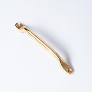 May include: A gold-colored cabinet pull with a curved handle and two mounting points. The hardware is made of metal and has a smooth, polished finish. The pull is designed for use on drawers or cabinet doors.
