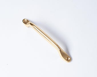 Op de afbeelding: Een goudkleurige kastgreep met een gebogen handvat en twee bevestigingspunten. De metalen hardware heeft een gladde, gepolijste afwerking. De greep is ontworpen voor gebruik op laden of kastdeuren. Ongeveer 15 cm lang.
