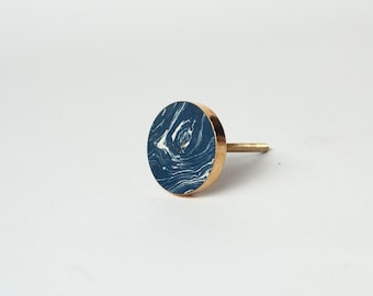 Op de afbeelding: Een ronde meubelknop met een donkerblauw oppervlak met witte gemarmerde wervelingen. De knop heeft een metalen goudkleurige zijkant en een metalen schroefdraadbout die aan de achterkant uitsteekt voor bevestiging op meubels of kastdeuren.