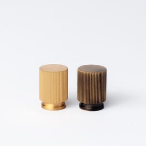 May include: Two cylindrical cabinet knobs. One is a light gold colour, and the other is a darker bronze. Both knobs have vertical ribbed detailing on the sides and a flat top. The knobs are set against a white background.