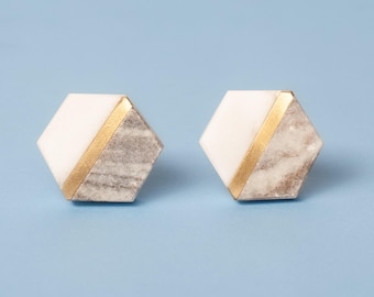 Op de afbeelding: Een paar zeshoekige oorknopjes met een gespleten ontwerp. De ene helft van elke oorbel is effen wit, terwijl de andere helft een grijs en wit marmerpatroon heeft. Een dunne, diagonale goudkleurige metalen band scheidt de twee secties.