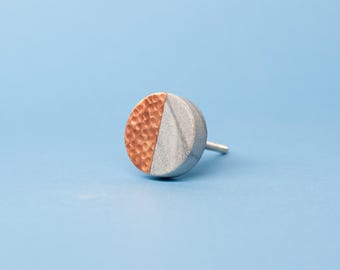 Op de afbeelding: Een ronde meubelknop met een gespleten ontwerp, waarbij de ene helft een textuur van gehamerd koper heeft en de andere helft een gladde, matte grijze afwerking, compleet met een metalen bevestigingspen die aan de achterkant uitsteekt.