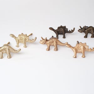 May include: A collection of five dinosaur-shaped cabinet knobs made of metal. The set includes three triceratops and two stegosaurus figures in varying metallic finishes, including polished gold and antique bronze tones, each with a mounting screw.