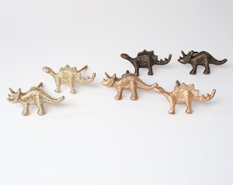 Op de afbeelding: Een set van vijf dinosaurusvormige kastknoppen gemaakt van metaal. De collectie bevat drie triceratops- en twee stegosaurus-figuren in verschillende metallic afwerkingen, waaronder gepolijst goud en antiek brons, elk voorzien van een schroef.
