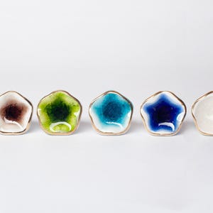 May include: Five small, star-shaped ceramic dishes with gold rims. The dishes are in a row and feature brown, green, turquoise, blue, and white colours. The dishes are set against a white background.