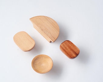 Op de afbeelding: Een set van vier houten meubelknoppen in verschillende vormen en houttinten. De set bevat een grote halfronde knop, twee kleinere ovale knoppen in licht- en middenbruin hout, en één kleine ronde knop met een holle, concave vorm in het midden.