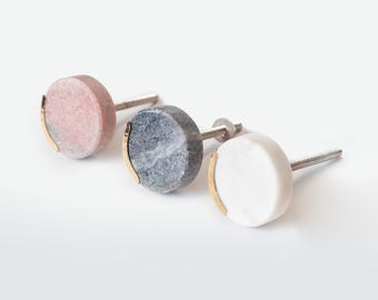 Op de afbeelding: Drie ronde kastknoppen met een glad stenen oppervlak in roze, grijs en wit. Elke knop is aan één kant voorzien van een dunne, gebogen goudkleurige metalen rand en heeft een metalen bout met schroefdraad aan de achterkant voor montage.