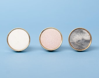 Op de afbeelding: Drie ronde kastknoppen met een goudkleurige metalen rand. De linkerknop heeft een inleg van effen witte steen, de middelste knop heeft een inleg van lichtroze steen en de rechterknop toont een grijs en wit gemarmerd steenpatroon.