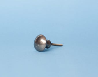 Op de afbeelding: Een enkele, donkergrijze kastknop met een afgeronde, ovale vorm. De knop heeft een metallic afwerking en een schroefdraad voor bevestiging. De achtergrond is effen lichtblauw.