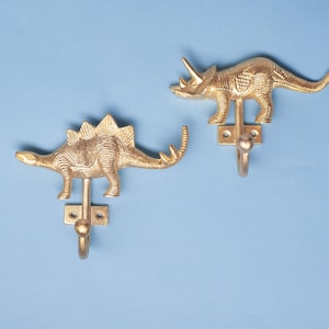Könnte beinhalten: Zwei goldfarbene Wandhaken aus Metall in Dinosaurierform. Ein Haken zeigt einen Stegosaurus mit Platten auf dem Rücken, der andere einen Triceratops mit drei Hörnern. Jeder Haken verfügt über eine rechteckige Montageplatte mit zwei Schraublöchern.