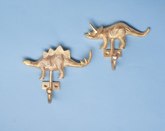 Op de afbeelding: Twee goudkleurige metalen wandhaken in de vorm van dinosaurussen. De ene haak toont een Stegosaurus met platen op de rug, de andere een Triceratops met drie hoorns. Elke haak heeft een rechthoekige montageplaat met twee schroefgaten.