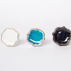 May include: Three ceramic cabinet knobs in white, turquoise, and black. Each knob has a unique, organic shape with a gold-coloured rim. The knobs are designed for furniture and home decor, offering a stylish accent.