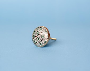 Op de afbeelding: Een ronde meubelknop met een glanzend oppervlak voorzien van een herhalend patroon van groene en witte sterren op een zwart-witte geometrische achtergrond, afgewerkt met een goudkleurige metalen voet en een korte bevestigingsschroef.