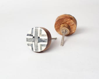 Op de afbeelding: Twee houten kastknoppen. Eén knop heeft een cirkelvormig ontwerp met een kruispatroon in grijstinten en wit. De andere knop is van hout. Beide knoppen hebben een metalen schroef voor installatie.