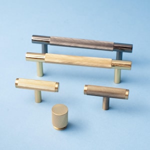 May include: A selection of cabinet handles and knobs with a knurled texture. The hardware includes T-bar handles and a cylindrical knob, in gold, bronze, and gunmetal finishes. The items are displayed against a light blue backdrop.