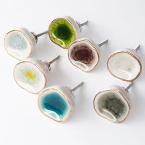 May include: A set of seven decorative cabinet knobs. Each knob has a unique, colourful, geode-like design with shades of green, blue, brown, and yellow. The knobs have a silver stem and a gold-coloured trim.