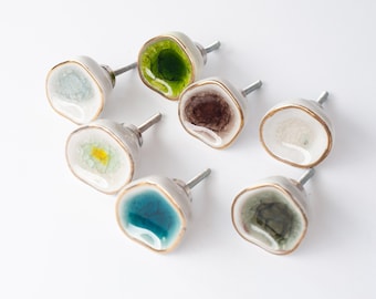 Op de afbeelding: Een verzameling van zeven decoratieve kastknoppen. Elke knop heeft een uniek, kleurrijk, geode-achtig ontwerp met tinten groen, blauw, bruin en geel. De knoppen hebben een zilveren steel en een goudkleurige rand.