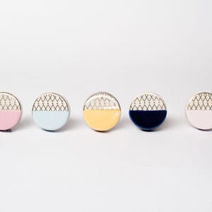May include: Five decorative cabinet knobs in pastel shades: pink, pale blue, yellow, navy, and pink. Each knob features a silver edge and a geometric design on the top half. The lower half of each knob is a solid colour.