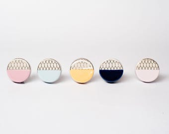 Op de afbeelding: Vijf ronde kastknoppen in pastelkleuren: roze, lichtblauw, geel, marineblauw en roze. Elke knop heeft een zilveren rand en een geometrisch patroon op de bovenste helft. De onderste helft van elke knop is effen van kleur.