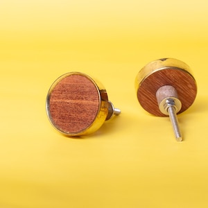 Può includere: Due pomelli decorativi per mobili. Ogni pomello presenta un centro rotondo in legno marrone-rossiccio, racchiuso in una cornice di metallo color oro. I pomelli sono posti su uno sfondo giallo brillante, che ne mette in risalto il design.