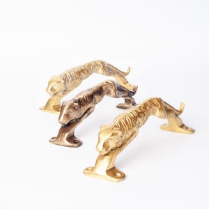 May include: Three metal door handles shaped like leaping tigers. Two handles have a bright gold-toned finish, while the center handle features a darker, antiqued bronze-toned finish. Each handle has mounting plates at the front and back paws.