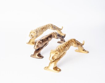 Op de afbeelding: Drie metalen deurgrepen in de vorm van springende tijgers. Twee grepen hebben een heldere goudkleurige afwerking, terwijl de middelste greep een donkerdere, antiek-bronskleurige afwerking heeft. Elke greep heeft montageplaten bij de poten.